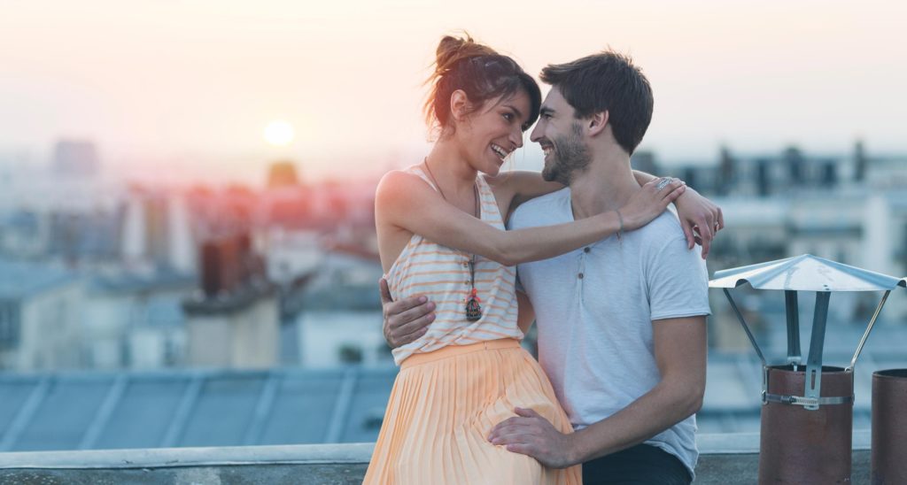Un couple assis sur un toit se prend dans les bras avec le couchée de soleil